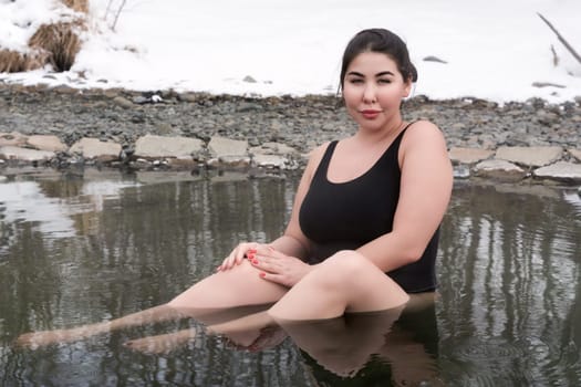 Outsize young adult woman in black one piece swimming costume sitting bathing in mineral water in outdoors pool at balneotherapy health spa. Femininity busty xxl woman looking at camera. Body positive