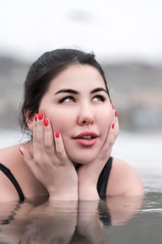 Woman embracing face with her palms, playfully looking up. Portrait of plus size brunette model in black swimming costume lying down in water of outdoors pool at balneotherapy spa, hot springs resort