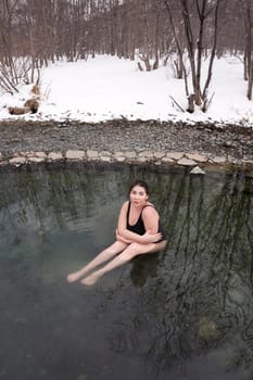 Large size young adult model in black one-piece bathing swimsuit sitting in geothermal mineral water in outdoors pool at balneotherapy spa, hot springs resort. Woman takes winter bathing and hardening