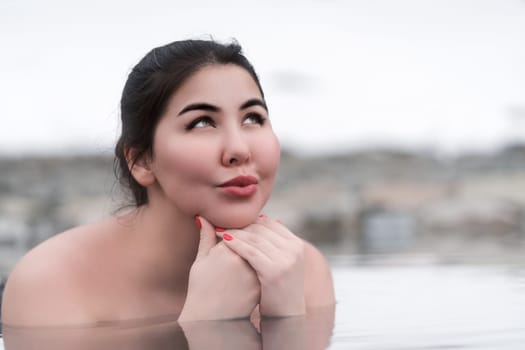 Portrait of femininity woman with folded palms under chin and looking up. Authentic plus size brunette model bathing and relaxation in mineral water of outdoor pool at balneotherapy spa. Body positive