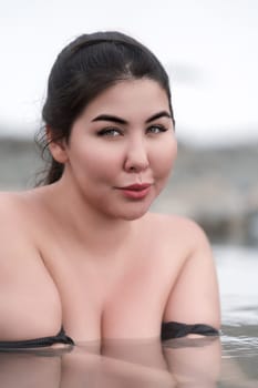 Portrait of plus size brunette woman with big breast bathing and relaxation in water of outdoor pool at balneotherapy health spa, hot springs resort. Busty full figured young model looking at camera