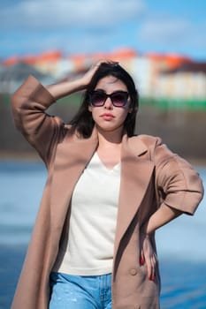 Portrait of stylish young woman with long hair, plump lips and sunglasses, dressed in beige coat, white sweater and blue jeans posing outdoors against background of blue lake and real estate