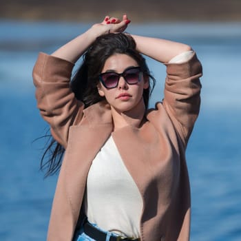Portrait of young woman with long hair, plump lips and sunglasses, dressed in beige coat and sweater posing outdoors against background of blue lake. Female looking at camera, hands raised above head