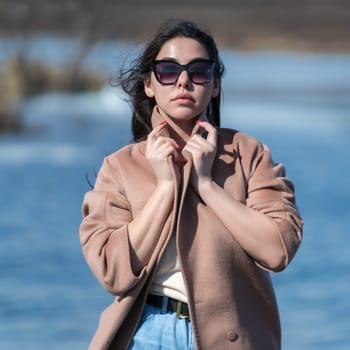 Glamorous hipster young woman in sunglasses, dressed in beige coat standing outdoors against background of blue lake on sunny spring day in good weather. Beauty female model looking at camera