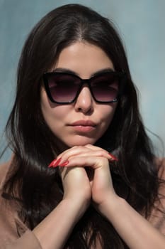 Glamorous young woman face with plump lips and glasses folded hands under her chin. Portrait of sexy hipster with long hair looks thoughtfully at camera through dark sunglasses.