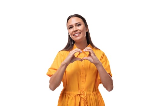 young woman dressed in a summer yellow dress shows a heart with her hands on a white background.