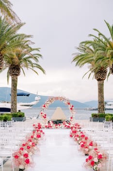Rows of chairs line a white path decorated with red flowers in front of a round wedding arch on the pier. High quality photo