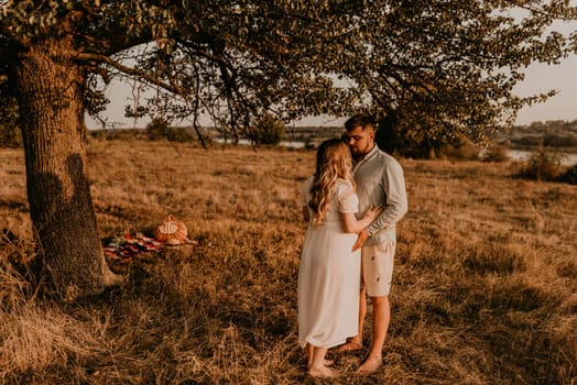 husband hugs pregnant wife with big tummy. Happy family resting in nature hugs kisses in summer at sunset. couple on background of large lonely tree. future mother Caucasian woman in cotton dress