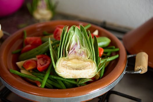 Selective focus on a half of ripe organic green artichoke and vegetables on the clay dish while cooking traditional Moroccan tagine. Food background