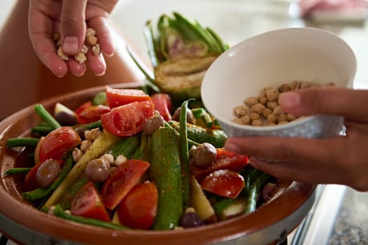 Closeup of the hands of a housewife adding chickpeas into a vegetarian meal while cooking healthy organic veggies in clay tagine pot. Traditional Moroccan food background