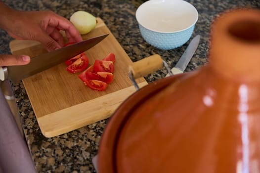 View from above of housewife chopping fresh organic ripe tomatoes on a wooden board while cooking dinner at home. Blurred clay tagine dish on the foreground. Food and healthy eating concept