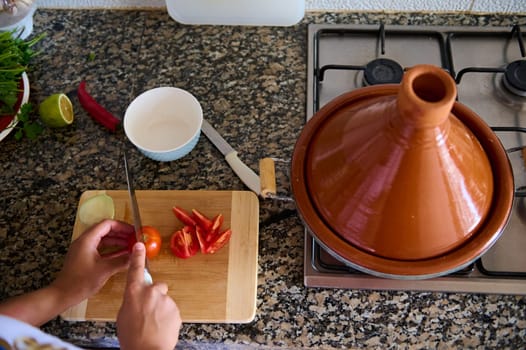 Overhead view of a woman housewife, standing at marble kitchen counter, slicing red juicy organic tomatoes, preparing salad, sauce or adding tomatoes into a vegetarian meal steaming in tagine clay pot