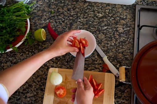 View from above of a chef slicing vegetables, adding chopped tomatoes into a white salad bowl while preparing dinner at home