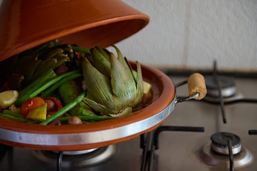 Cropped view of clay pot tagine with open loid while vegetables are steaming on the kitchen stove. Traditional Moroccan food concept