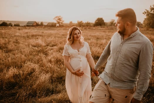 Happy family pregnant caucasian blonde woman with moles on face in white cotton dress walks with husband meadow summer. man in light natural clothes and shorts holds hand wife. trees in sunbeams