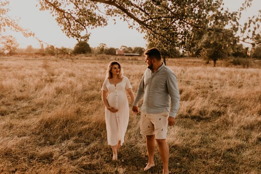 Happy family pregnant caucasian blonde woman with moles on face in white cotton dress walks with husband meadow summer. man in light natural clothes and shorts holds hand wife. trees in sunbeams