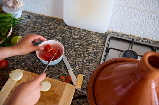 Top view housewife slicing fresh ripe tomatoes and onions on a cutting board, cooking healthy raw vegan salad. Ingredients lying on a marble kitchen counter and clay tajine pot on a steel stove