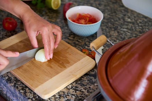 Cropped view of the hands of a chef cutting onion in two halves on a wooden board, using a kitchen knife. Housewife prepares dinner at home kitchen. Cuisine. Culinary. Domestic life