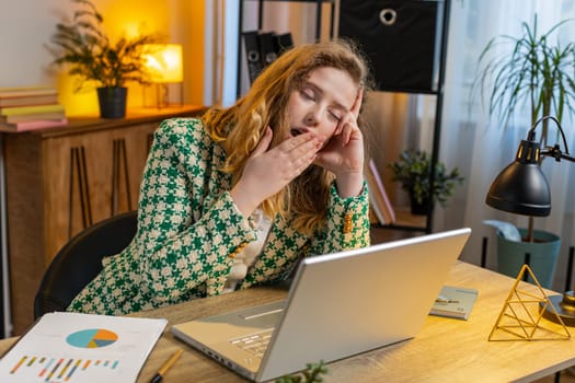 Bored sleepy young blond businesswoman worker working on laptop computer, yawning, leaning on hand at home office desk. Exhausted tired freelancer workaholic girl. Employment, occupation, workless.