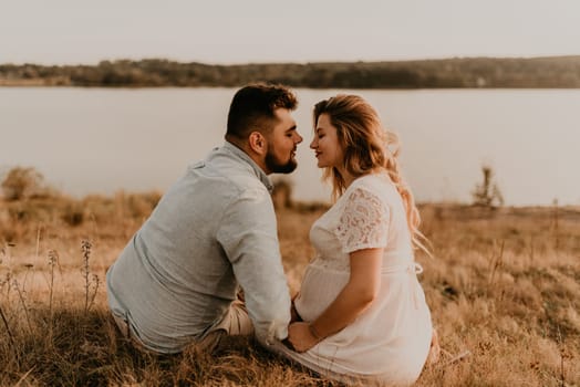 husband with pregnant wife with big belly tummy is resting on bank of river sitting on grass in summer sunny weather. couple in love caucasian man and woman hugging holding hands kissing outdoors
