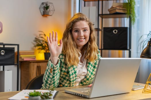 Hello. Happy Caucasian businesswoman employee waves hand hi gesture greeting welcomes someone, online webinar, remote distance conversation. Freelance girl sitting at desk using laptop in home office.