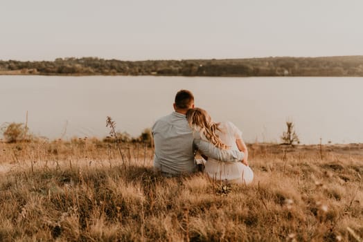 husband with pregnant wife with big belly tummy is resting on bank of river sitting on grass in summer sunny weather. couple in love caucasian man and woman hugging holding hands kissing outdoors