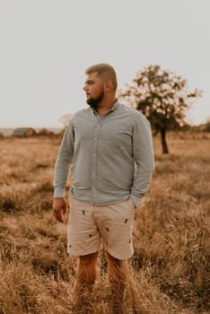 young caucasian black-haired overweight man with beard smiling cheerfully in summer cotton clothes and shorts outdoor in sunny weather. men fashion collection. open spaces meadow flooded sunlight tree