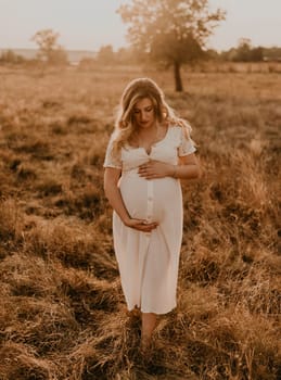 Caucasian pregnant young blonde woman in cotton white linen dress stand walking meadow on dry grass in summer at sunset nature. mother-to-be holds her hands on big round belly. tree in sunbeam