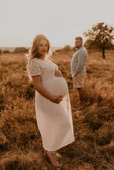 Happy family pregnant caucasian blonde woman with moles on face white cotton dress walks with husband in meadow in summer. man in light natural clothes and shorts holds hand wife. trees sunbeams