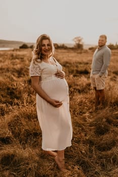 Happy family pregnant caucasian blonde woman with moles in white cotton dress walks with husband in meadow in summer. man light natural clothes and shorts holds hand wife. trees sunbeams