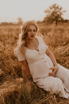 Caucasian pregnant young blonde woman in cotton white linen dress sits and lies in the middle of the meadow on dry grass in summer at sunset in nature. mother-to-be holds her hands on big round belly