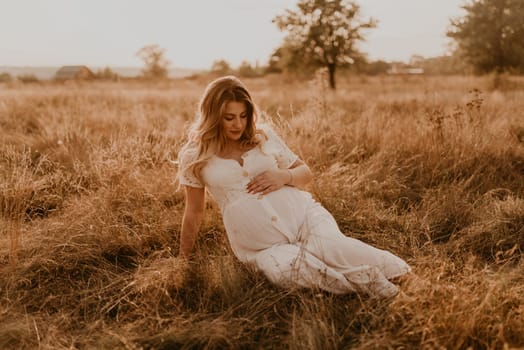 Caucasian pregnant young blonde woman in cotton white linen dress sits and lies in the middle of the meadow on dry grass in summer at sunset in nature. mother-to-be holds her hands on big round belly