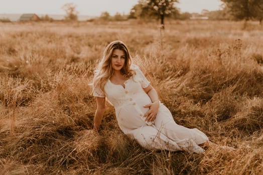 Caucasian pregnant young blonde woman in cotton white linen dress sits and lies in the middle of the meadow on dry grass in summer at sunset in nature. mother-to-be holds her hands on big round belly