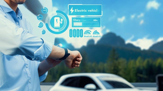 Businessman checking EV car digital battery status in hologram from smartwatch recharging at charging station, vacation and travel using eco-friendly electric car in natural outdoor background. Peruse