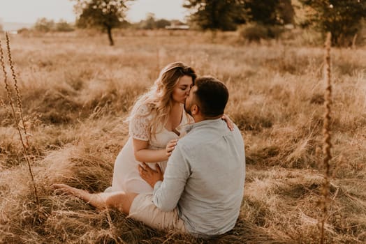 pregnant wife sits in her husband arms and kisses and hugs him. Happy family resting nature hugs kisses in summer at sunset. future mother Caucasian woman white cotton dress father in shorts