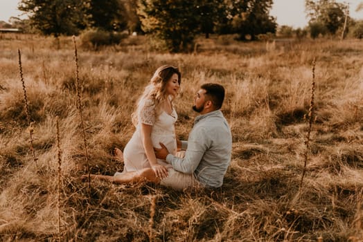 pregnant wife sits in her husband arms and kisses and hugs him. Happy family resting nature hugs kisses in summer at sunset. future mother Caucasian woman white cotton dress father in shorts