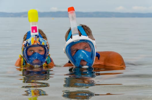 Child and an adult wearing snorkeling masks. Only their heads are visible above water. Faces in masks are reflected in water. Sea and sky are blue all around. Space for text.