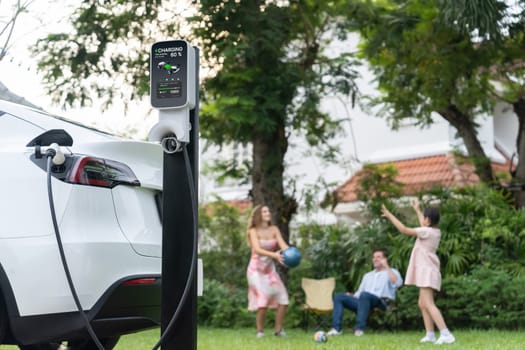 Focus electric vehicle recharge from home charging station on blur background of happy and playful family playing together. EV car using alternative and sustainable energy for better future.Synchronos