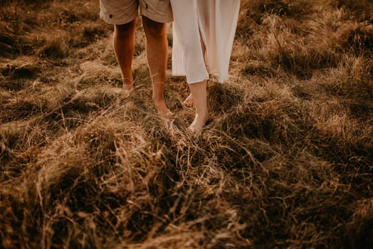 tanned man and light skin legs woman barefoot close-up walking on dry green thin grass. couple in love in summer in thin light cotton clothes walks in hot warm weather. women dress and men shorts