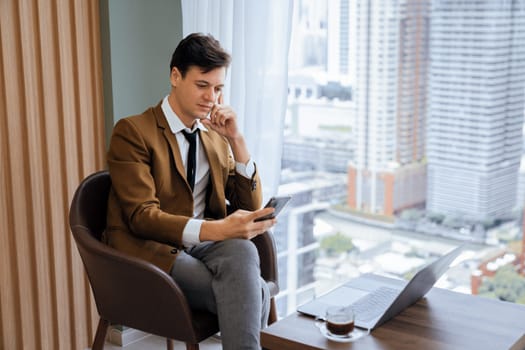 Closeup of handsome businessman using mobile phone while sitting near window with skyscraper view. Professional male leader taking a photo marketing, business project displayed on laptop. Ornamented.