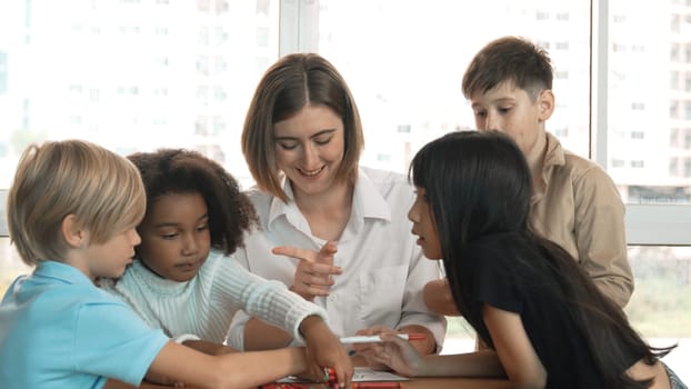 Professional caucasian teacher telling story to diverse student while sitting at table with storybook and colored book. Smart learner listening story while colored picture from instructor. Erudition.