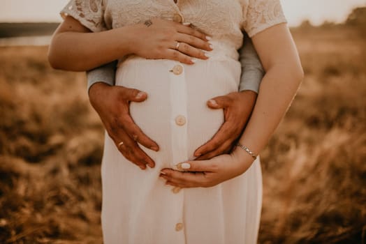 hands of future mother and father clasped pregnant big tummy. husband hugs pregnant wife. Happy family resting in nature hugs in summer at sunset. Caucasian woman in white cotton dress