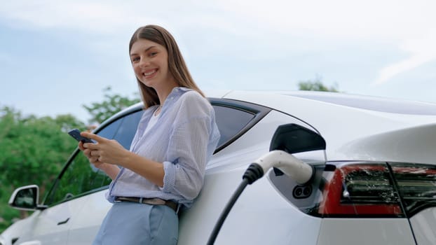 Young woman recharging battery for electric car during road trip travel EV car in natural forest or national park. Eco friendly travel during vacation and holiday. Exalt