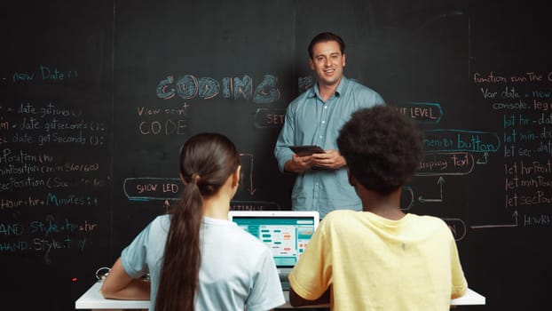 Caucasian teacher answering student question while holding tablet. Caucasian girl pointing at blackboard with code or prompt while laptop display system screen at STEM technology class. Edification.