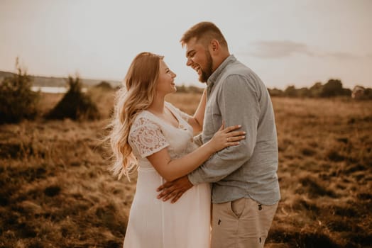 husband laughs hugs pregnant wife keep his hands on big round tummy. Happy family resting in nature hugs kisses in summer at sunset. future mother Caucasian woman in white cotton dress father