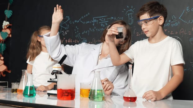 Caucasian boy mixing chemical liquid while teacher giving advice. Professional instructor wearing lab suit looking for diverse student at table with beaker filled with colored solution. Erudition.