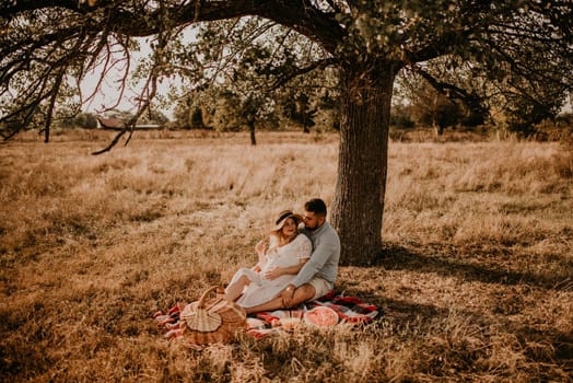 happy European caucasian family with a pregnant hug laugh having fun. expectant mother in hat and dress in summer under tree on bedspread.