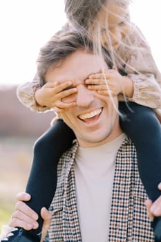 Little girl covered her laughing dad eyes with her palms while sitting on his shoulders. High quality photo