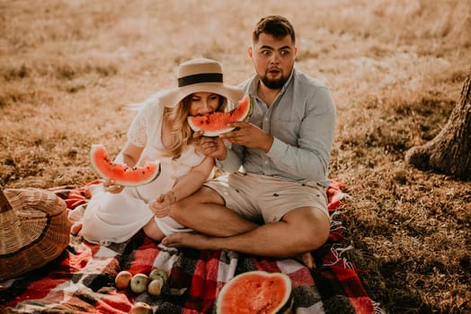 happy European caucasian family with a pregnant woman relaxing in nature picnic eating fruit red juicy watermelon laugh having fun. expectant mother in hat and dress eating watermelon in summer.