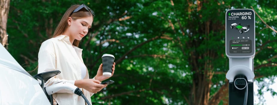 Young woman with coffee and sustainable urban commute by EV electric car recharging at outdoor cafe in springtime garden, green city sustainability and environmental friendly EV car.Panorama Expedient